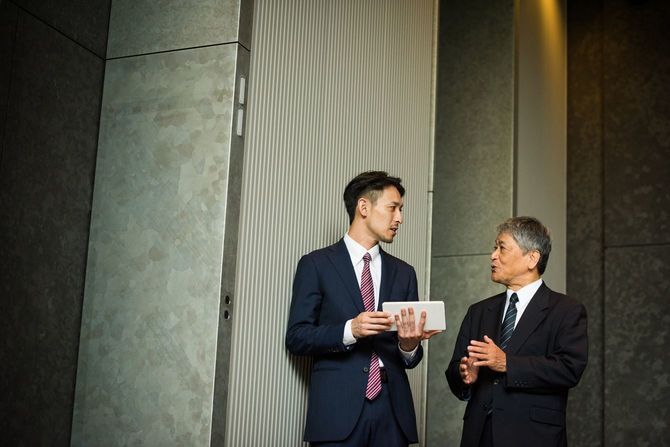 オフィスのエレベーターホールで立ち話をする二人のビジネスパーソン
