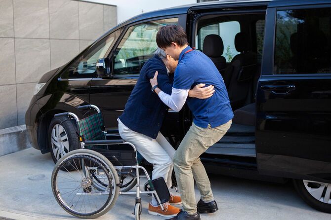 若い男性介護者に助けられて車椅子から車に乗り込む老人