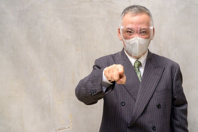険しい表情でこちらを指さすマスクとフェイスシールドをつけたスーツの中年男性