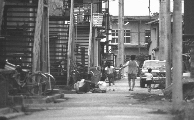 路地を行く手をつないだ3人の子供を座って見ている子供たち