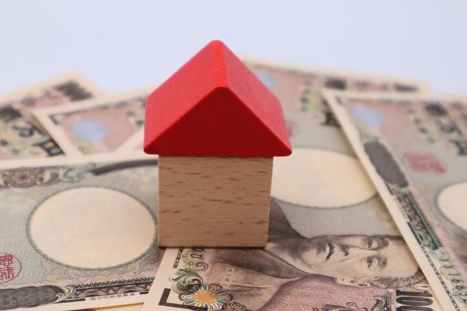 散らばった一万円札の上に置かれた家の形の木製フィギュア