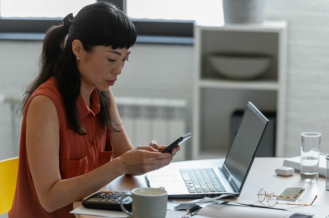 自宅オフィスでノートパソコンを使いながら、スマホを見る女性