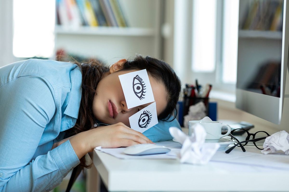 目の絵が描かれた紙を目の辺りに貼って机で寝ている女性